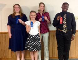 White girl in dark blue dress holding a blue ribbon, white girl with white blouse and black and white skirt holding a blue ribbon, white girl with dark red top and khaki pants holding a blue ribbon and black boy with black shirt, striped tie and black slacks holding a red ribbon.